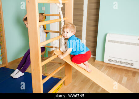 Süße kleine Jungen barfuß in einem blauen T-Shirt klettern auf die hölzerne Folie zu Hause sport Komplex. Kinder spielen auf einer Holzleiter Stockfoto