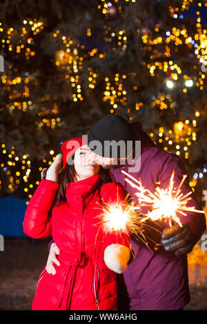 Portrait von süßen küssen paar in warme Kleidung, mit funkelnden Bengal Leuchten in ihren Händen, das stehen unter eingerichtet Stadt Weihnachtsbaum. Happy lif Stockfoto