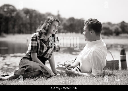 1940 lächelnde junge Paar MANN UND FRAU SITZEN liegen auf Gras von Teich sich EINANDER genießen ein Picknick-c 3414 HAR 001 HARS NOSTALGIE OLD FASHION 1 KOMMUNIKATION junger Erwachsener BALANCE TEICH TEAMARBEIT STARKE ENTSPANNENDE FREUDE FREUDE LIFESTYLE FRAUEN VERHEIRATET LÄNDLICHEN EHEPARTNER EHEMÄNNER GESUNDHEIT KOPIE RAUM FREUNDSCHAFT IN VOLLER LÄNGE mit halber Länge DAMEN FITNESS PERSONEN INSPIRATION FÜRSORGLICHE MÄNNER B&W PARTNER DATING WEITWINKEL TRÄUME GLÜCK FRÖHLICHE FREIZEIT KRAFT UND WAHL GENIESSEN AUFREGUNG ERHOLUNG AN, INDEM SIE AUF ANZIEHUNG LÄCHELT VERBINDUNG UMWERBUNG KONZEPTIONELLE ESCAPE FREUDIGE STILVOLLE Stockfoto