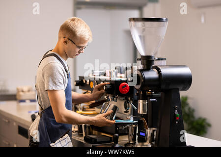 Junge männliche Barista in Uniform mit Stahl Becher stehen moderne Kaffeemaschine Stockfoto