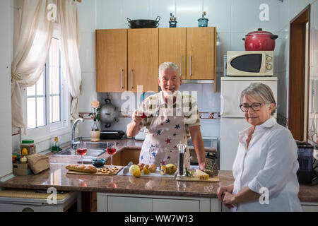 Ein erwachsener älterer Mann Wein zu trinken und dabei etwas für einen Cocktail mit ihrer Frau zu Essen vorbereiten. Altenheim arbeiten in einem üblichen Küche. Licht vom Fenster. Stockfoto