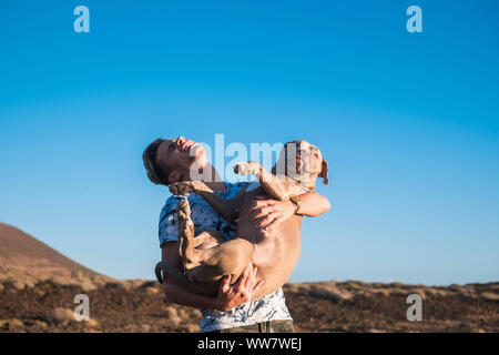 Junge blonde schöne Mann und seinen geliebten Hund mit viel Spaß zusammen Lachen und Lächeln in Outdoor Freizeitaktivitäten Aktivität. Der junge Umarmung und das Tier der beste Freund durchführen Stockfoto