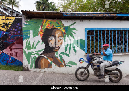 Mann auf dem Motorrad fahren vor der Wand eines Hauses mit bunten Graffiti von einer Frau Stockfoto