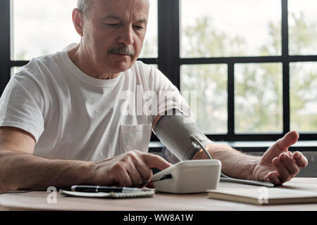 Alte Hispanic Mann schlechte Gefühl mit einem Haus Blutdruck Maschine seine Gesundheit am Morgen zu prüfen. Stockfoto