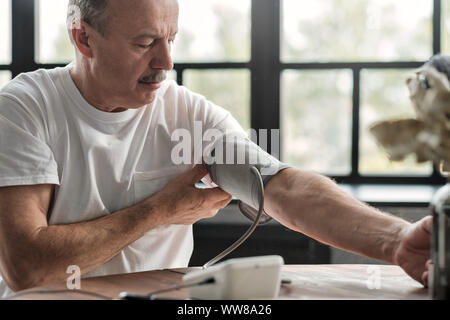 Alte Hispanic Mann schlechte Gefühl mit einem Haus Blutdruck Maschine seine Gesundheit am Morgen zu prüfen. Stockfoto