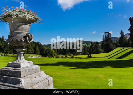 Eine reich verzierte Topf mit Blumen und breiten grünen Rasen in Powerscourt Gärten, Wald und Sugerloaf Berg im Hintergrund, Wicklow, Irland Stockfoto