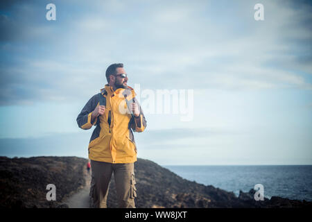 Männliche Trekker wandern und genießen die Natur an der Küste in einer Vulkaninsel. Blick auf das Meer und die Wüste um ihn herum. allein Reisen und Orte Konzept entdecken. blau Töne für kalten Tag Stockfoto