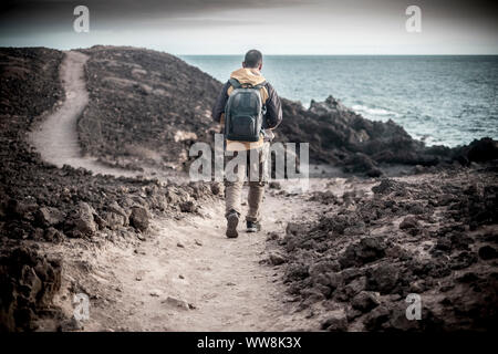 Einsamen Reisenden jungen Mann mittleren Alters mit Trekking Kleidung und Rucksack wandern auf dem Ozean Küste in einem Felsen Wüste. Nach einem Pfad mit Meerblick und genießen Freiheit und alternativer Lebensstil Stockfoto