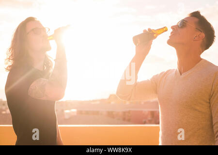 Ein paar junge Männer trinken Bier zusammen unter den Sonnenuntergang Sonnenlicht. helles Bild für schöne Männer Modelle in Glück und Outdoor Freizeitaktivitäten Aktivität. Auf der Dachterrasse mit Blick auf die Stadt Stockfoto