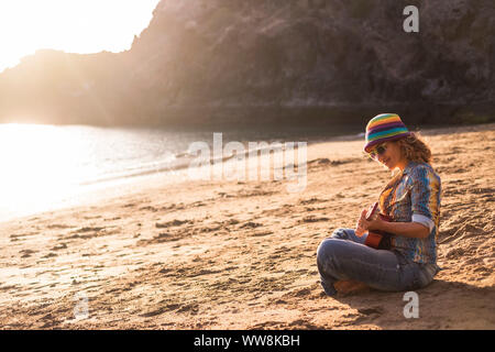 Schöne und ruhige junge Frau sitzt am Ufer am Strand anjoying Freizeitaktivitäten Aktivität auf dem Sand spielen eine ukulele Gitarre in der Nähe des Ozeans für Urlaub und Freiheit Konzept lächelnd Stockfoto