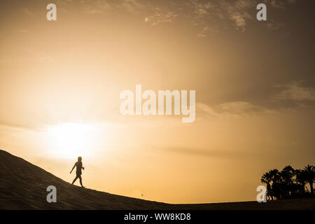 Dame zu Fuß auf einer Düne Wüste in Maspalomas Kanarische Inseln während eines goldenen smazing schönen Sonnenuntergang mit orangen und gelben Farben. tropische Palmen im Hintergrund, alleine in der Natur ocncept Stockfoto