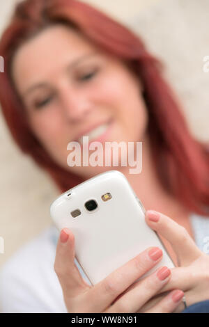 Hübsches Lächeln rothaarige Frau, die das Senden von Nachrichten oder ein selfie auf Ihr Handy in einem Park machen. Konzentrieren Sie sich auf Ihr Telefon Stockfoto