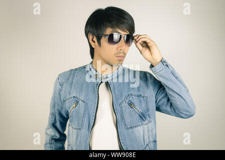 Jungen asiatischen Mann in Jeans Jacke Jeansjacke Berühren oder Sonnenbrille. Denim oder Jeans Jacket Men Fashion auf grauem Hintergrund Stockfoto