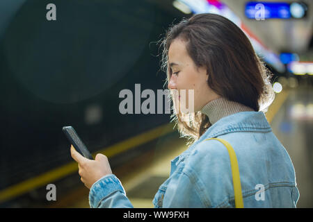 Jugendlicher Mädchen in Jeans mit Rucksack auf Metro Station Holding smart Telefon in der Hand, Blättern und texting, zu lächeln und zu lachen. Finnland Stockfoto