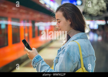 Jugendlicher Mädchen in Jeans mit Rucksack auf Metro Station Holding smart Telefon in der Hand, Blättern und texting, zu lächeln und zu lachen. Finnland Stockfoto