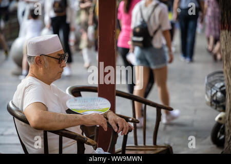 Xian, China - Juli 2019: Müde Chinesischer Mann ruht auf einem Stuhl und selbst die Kühlung durch Winken mit einem kleinen Ventilator an einem heißen Tag auf der Straße in der MUSLI Stockfoto