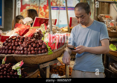 Xian, China - Juli 2019: gelangweilt Obst Verkäufer sein Smartphone Kontrolle auf einem im muslimischen Viertel Abschaltdruck Stockfoto