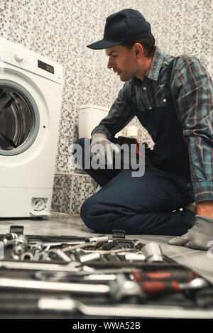 Porträt der jungen Handwerker in Uniform neben gebrochenen Waschmaschine und versuchen, es zu beheben. Vertikale erschossen. Seitenansicht Stockfoto