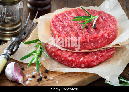 Rohes Fleisch Rindfleisch Burger auf einem hölzernen Schneidebrett mit frischem Rosmarin hacken, Knoblauchzehen, Pfefferkörner und Salz Stockfoto