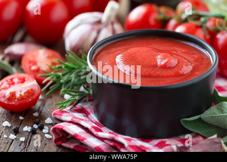 Tomatensoße oder Ketchup in einem schwarzen Schüssel mit Zutaten zum Kochen im Hintergrund Stockfoto