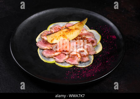 Kantige Stücke mit karamellisierten Birnen auf einem schwarzen Keramik Teller. Gebackenes Fleisch in dünne Stücke geschnitten Stockfoto