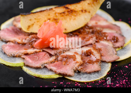 Kantige Stücke mit karamellisierten Birnen auf einem schwarzen Keramik Teller. Gebackenes Fleisch in dünne Stücke geschnitten Stockfoto