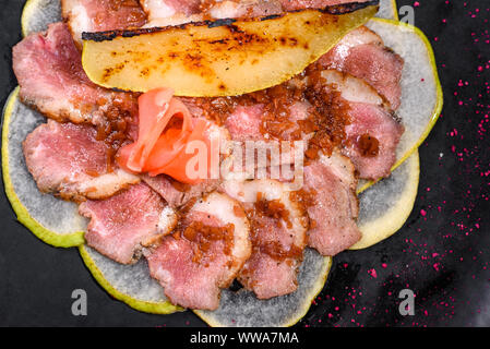 Kantige Stücke mit karamellisierten Birnen auf einem schwarzen Keramik Teller. Gebackenes Fleisch in dünne Stücke geschnitten Stockfoto