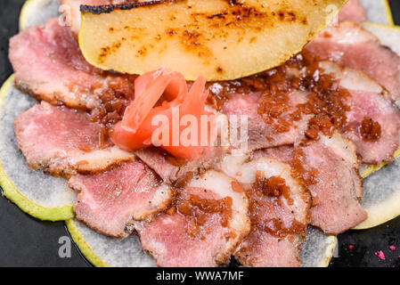 Kantige Stücke mit karamellisierten Birnen auf einem schwarzen Keramik Teller. Gebackenes Fleisch in dünne Stücke geschnitten Stockfoto