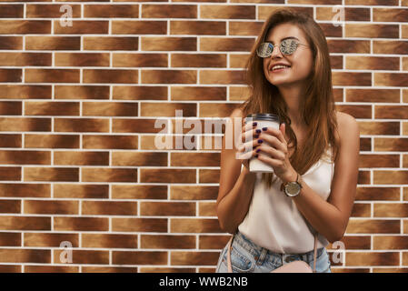 Portrait von fröhliche junge Frau mit Sonnenbrille über Ziegel Hintergrund stehen während outdoor Fotoshooting. Sie ist weg schauen mit einem Lächeln, während Sie Ihr Essen trinken halten. Horizontale Schuß Stockfoto