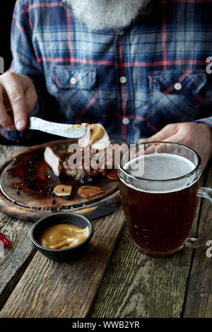 Nahaufnahme der brutalen Grauhaarige erwachsenen Mann mit einem Bart Senf Steak isst und trinkt Bier, das Konzept eines Urlaubs, Festival, Oktoberfest oder St. Patrick's Da Stockfoto