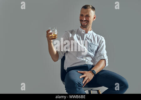 Mann mit Schnurrbart, in klassischem Weiß Hemd und blauer Hose sitzt auf einem Stuhl, mit einem Glas Whiskey. Grauer Hintergrund, Nahaufnahme. Stockfoto