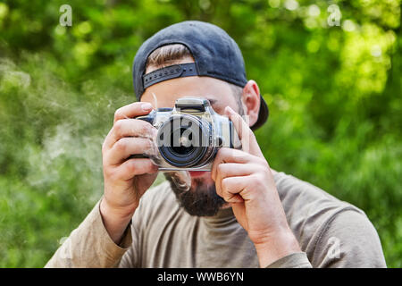 Die Kamera raucht in den Händen eines jungen Bartgeier Fotograf in einem Baseball Cap, Visor, der ein Foto in die Natur nimmt. Ein männlicher Photographe Stockfoto