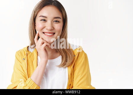 Close-up Fröhliche charismatische asiatische blonde Mädchen berühren Wange dumme Breit grinsend Schauen motiviert süße Grinsen toothy zufrieden Ausdruck genießen Sie reine Stockfoto