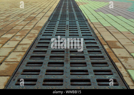 Quadratischer Abwasserkanal, strukturierter Boden neben einem Abfluss in der Mitte eines Parks nach Regen, Boden aus Blöcken und derzeit nass. Alte gestreifte Entwässerungsrinne Käfig Stockfoto