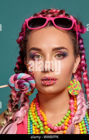 Schöne Mädchen mit einem bunten Zöpfe Frisur und Make-up, im Studio vor einem blauen Hintergrund posiert, Holding einen Lutscher in der Hand. Stockfoto