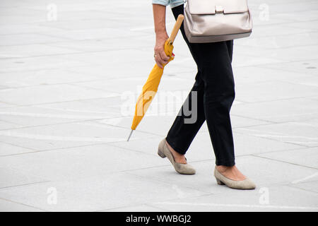 Regen: eine Frau zu Fuß auf Stadt squre Pflaster, die gefaltet yellow umbrella, von der Taille abwärts Stockfoto