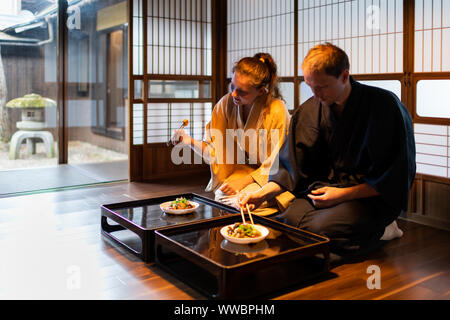 Paar im Kimono Seiza sitzen Holding Stäbchen, Essen in der traditionellen japanischen Ryokan Zimmer durch Tabelle Platten in der Nähe von Shoji Schiebetüren Türen Papier Stockfoto