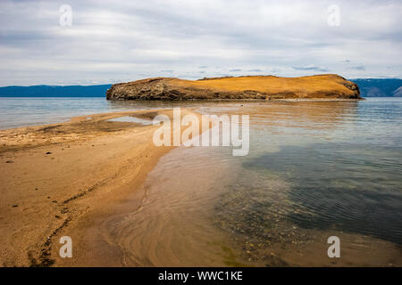 Eine Sandbank führt zu einer felsigen Insel am Baikalsee. Grauer Himmel, der in den Wolken. Algen und Kiesel sind sichtbar durch klares Wasser. Bergkette hinter t Stockfoto