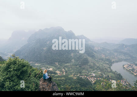 Junge kaukasier Frau steht oben auf dem Berg mit Blick auf das Flusstal in Nong Khiaw Dorf, Laos. Stockfoto