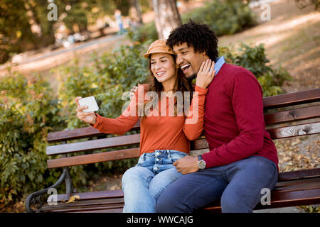 Multiratial junge Liebende Paar sitzt auf der Bank und unter selfie mit Handy im Herbst City Park Stockfoto