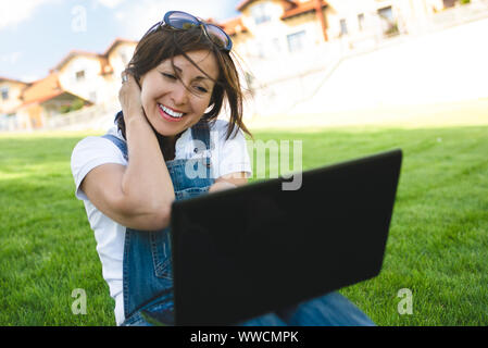 Weibliche erhält freudige Emotionen durch Gespräche mit ihren Freunden auf ein Konferenzgespräch aus Notebook, gekleidet in Jeans Overalls sitzen auf dem Rasen im Park Stockfoto