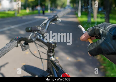 Eine Zigarette in der Hand eines rauchen Radfahrer, Fahrten mit dem Fahrrad auf dem Hintergrund der asphaltierte Weg des City Park Stockfoto