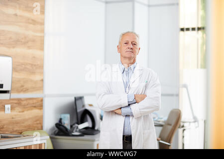 Portrait von erfahrenen Arzt tragen weiße Mantel in modernen Büro stand mit verschränkten Armen und Kamera posieren, kopieren Raum Stockfoto