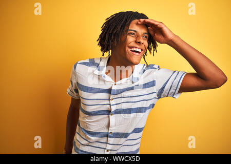 Afro-amerikanische Mann mit Dreadlocks tragen gestreifte Shirt über isolierte gelben Hintergrund sehr glücklich und lächelnd weit entfernt, mit der Hand über den Kopf. S Stockfoto