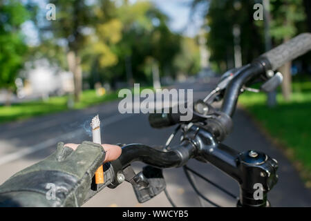 Eine Zigarette in der Hand eines rauchen Radfahrer, Fahrten mit dem Fahrrad auf dem Hintergrund der asphaltierte Weg des City Park Stockfoto