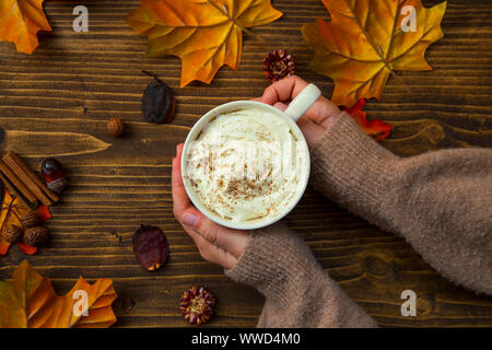 Herbst Kaffeetasse, Blick von oben auf die Frau, die Hände, die heiße Tasse Cappuccino mit Sahne und Zimt auf Holztisch mit getrockneten Blättern und autumna Stockfoto