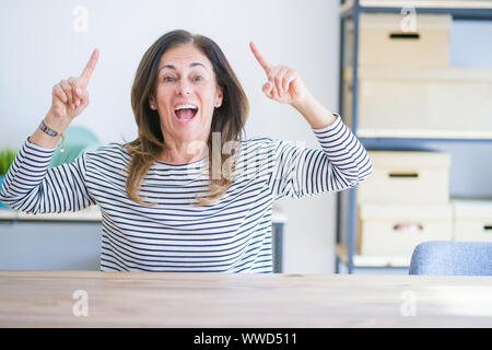 Mittleren Alters ältere Frau am Tisch zu Hause lächelnd erstaunt und überrascht und zeigt mit Fingern und erhobenen Arme sitzen. Stockfoto