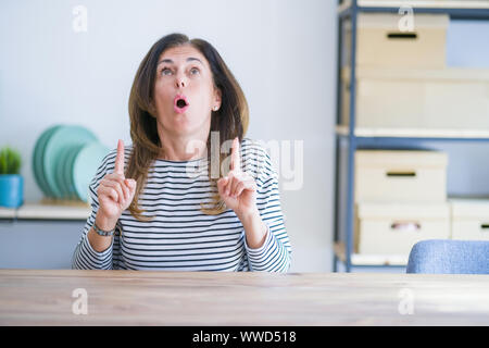 Mittleren Alters ältere Frau am Tisch sitzen zu Hause erstaunt und überrascht mit der Suche und zeigt mit Fingern und erhobenen Armen. Stockfoto