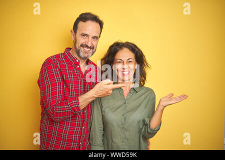 Schönen mittleren Alter Paar über isolierte gelben Hintergrund erstaunt und lächelt in die Kamera während der Präsentation mit Hand und zeigte mit dem Finger. Stockfoto