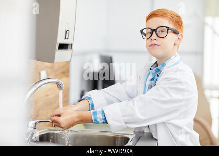 Taille bis Portrait von niedlichen rothaarigen Jungen tragen Laborkittel Händewaschen vor dem Verfahren im Doktorbüro, Kopie Raum Stockfoto